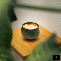 Durch ein Fenster aus grünen Blättern ist grün-schwarze Cappuccino Tasse mit Latte Art auf Holztisch.