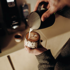 Person die Latte Art mit einem Milchkännchen in eine Cappuccino Tasse eingießt.