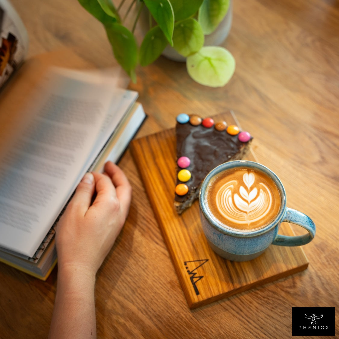 PHENIOX Cappuccino Tasse Ocean steht gefüllt mit einem Latte Art in Herzform auf einem Brettchen auf dem außerdem noch ein Stück Kuchen ist.