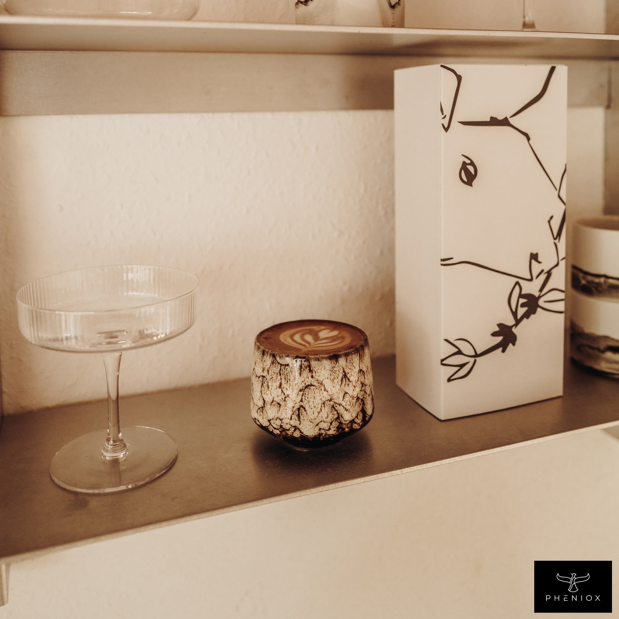 PHENIOX Cappuccino Tasse Tibet Weiß steht auf einem Regalbrett neben einem Cocktailglas und einer edlen Verpackung.