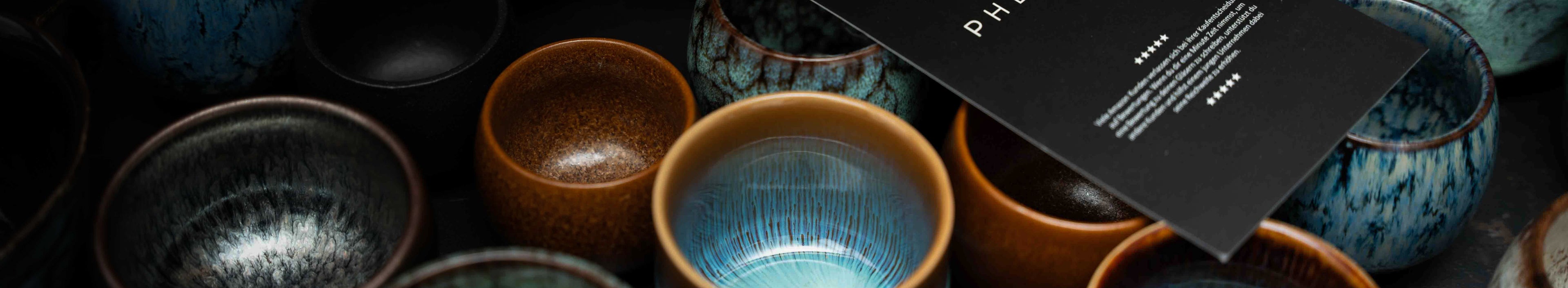 Viele Bunte PHENIOX Steinguttassen stehen nebeneinander. Auf ihnen liegt ein PHENIOX Flyer.